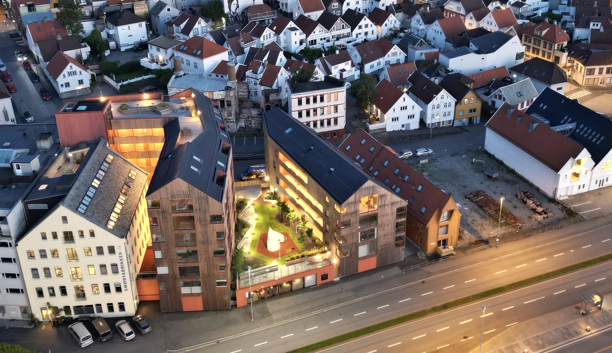 Flyfoto av Sameiet Skutene i blåtimen — to trekledde leilighetsbygg rundt et opplyst gårdsrom med Skroget-lekeapparatet, omkranset av Pedersgatas hvite trehus